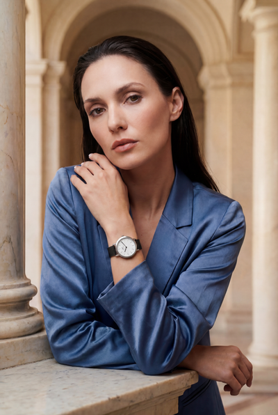 Woman in blue silk blazer wearing Aureum Swiss Made modular watch with round silver case and black mesh strap, posed by marble columns — Christopher Butendorff